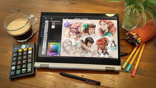 The photo shows the top of a wooden 
workbench with a foldable computer running Krita.

There is cup of coffee on the left and keypad for interacting with the art application. On the right you can see some brightly coloured pencils and a house plant in a glass.